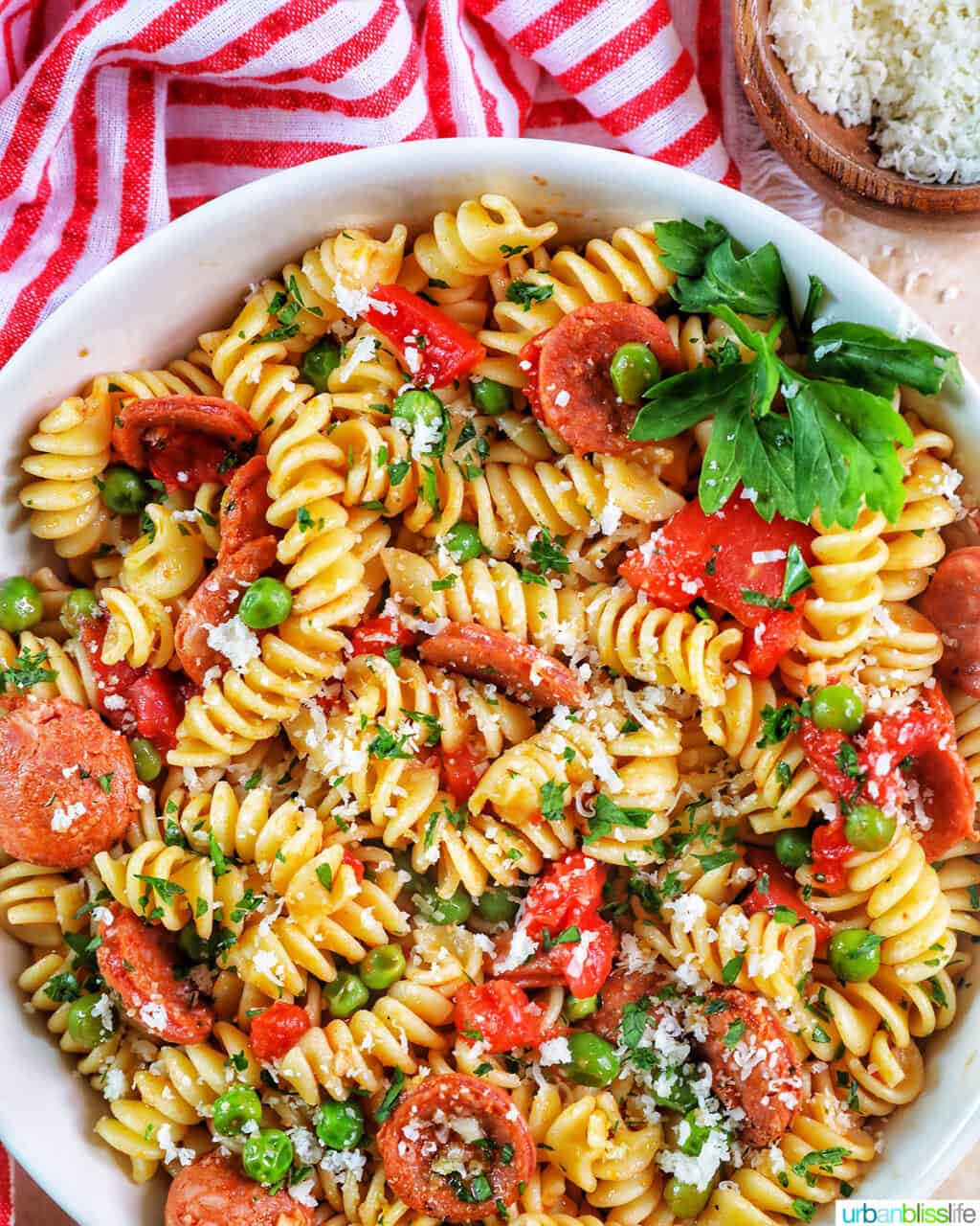 white bowl filled with pasta, tomatoes, herbs, peas, and linguica with red striped napkin..