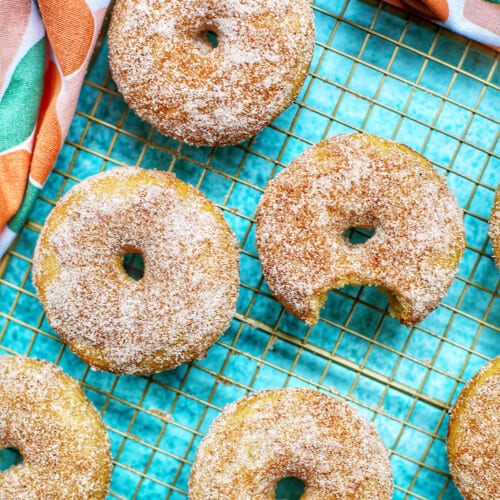 several apple cinnamon donuts on a wire rack, one with a bite taken out of it.