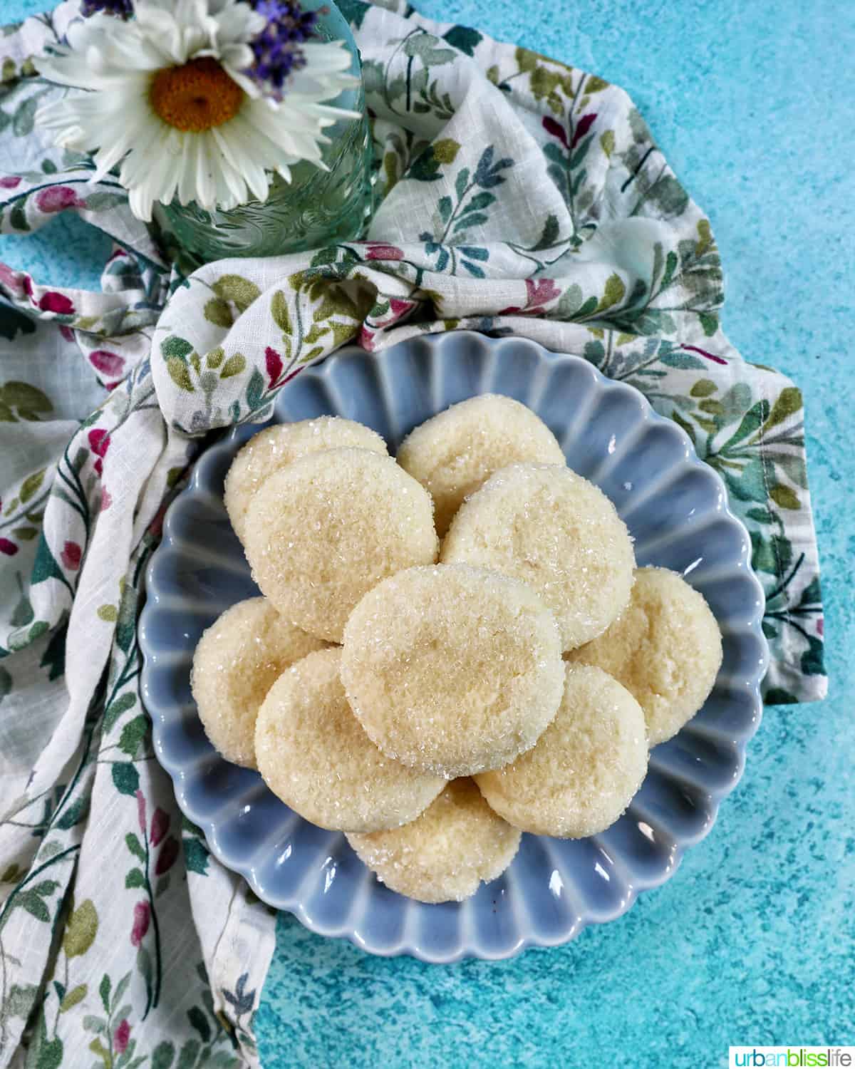blue plate with stacked soft almond sugar cookies on a colorful napkin on blue table, with daisy flowers.
