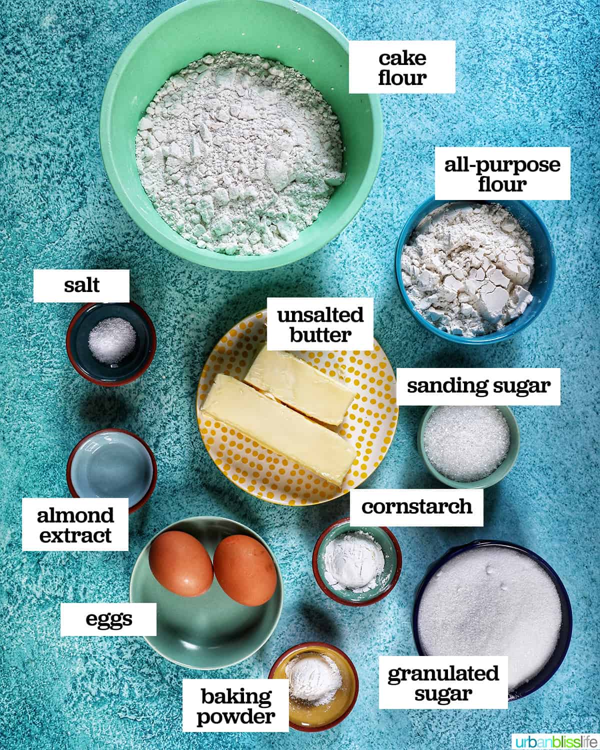 bowls of many ingredients to make soft almond sugar cookies on a blue background.