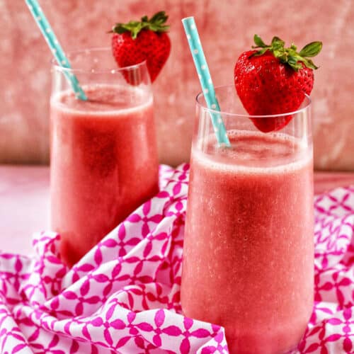 two glasses with strawberry watermelon smoothies, blue straws, sliced strawberry garnish on a pink and white napkin.