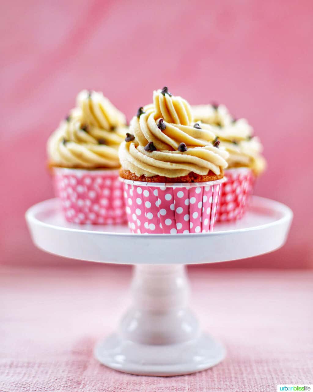 Three Chocolate Chip Cupcakes with Cookie Dough Frosting on a white cake pedestal with pink background