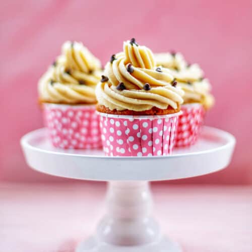 Three Chocolate Chip Cupcakes with Cookie Dough Frosting on a white cake pedestal with pink background