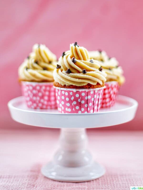 Three Chocolate Chip Cupcakes with Cookie Dough Frosting on a white cake pedestal with pink background