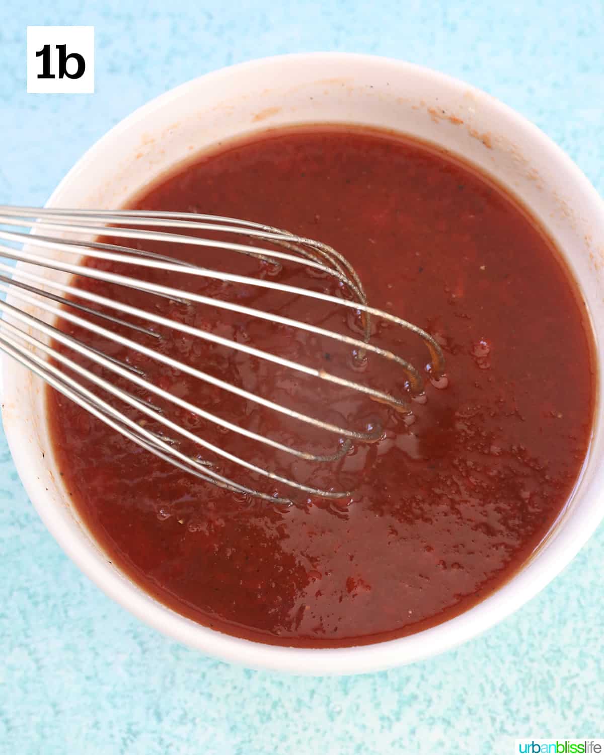 white bowl with sauce for honey bourbon meatballs and a whisk in the bowl, on top of a blue background.