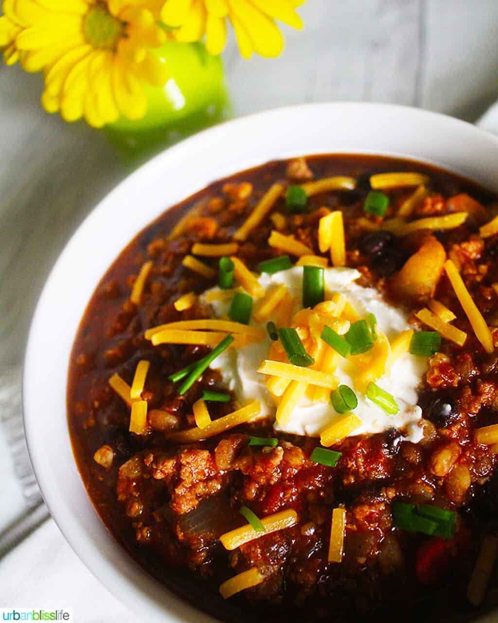 bowl of turkey chili with ground turkey, farro, stout, and toppings with a vase of yellow daisies.