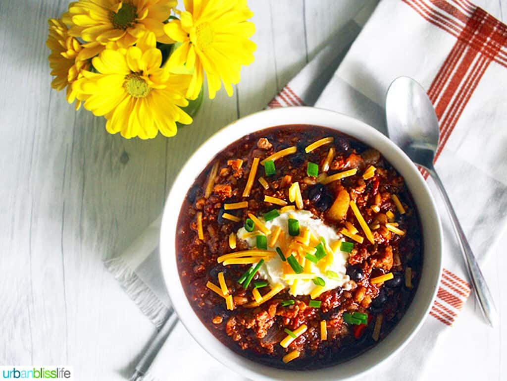 bowl of turkey chili with ground turkey, farro, stout, and toppings with a vase of yellow daisies.