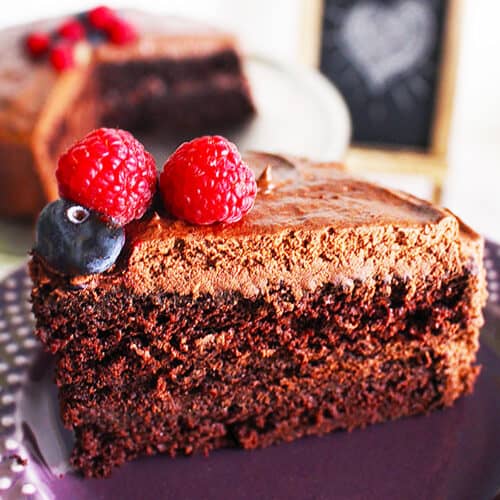 slice of vegan chocolate cake with chocolate buttercream frosting, fresh fruit on top of a purple plate.
