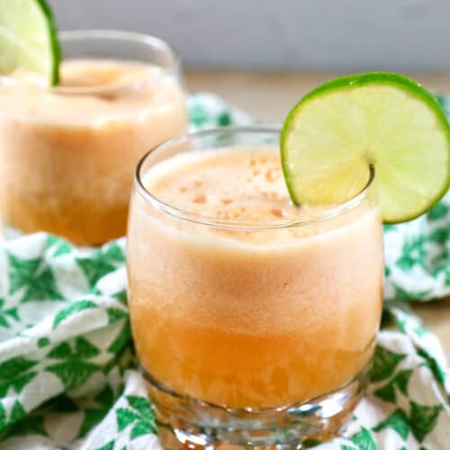 side view of two glasses of cantaloupe margaritas with lime slice garnish on a green and white patterned napkin.