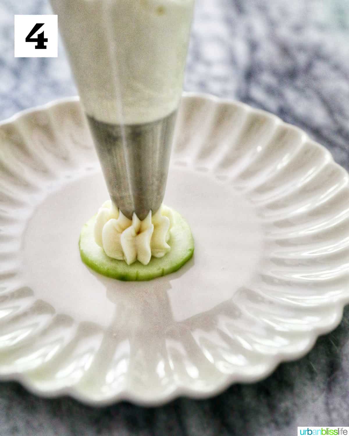 cream cheese piping out of a pastry bag onto a cucumber slice on a scalloped plate.
