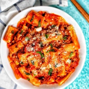 bowl of rigatoni pasta with Italian sausage on a white and blue checkered napkin and a fork.
