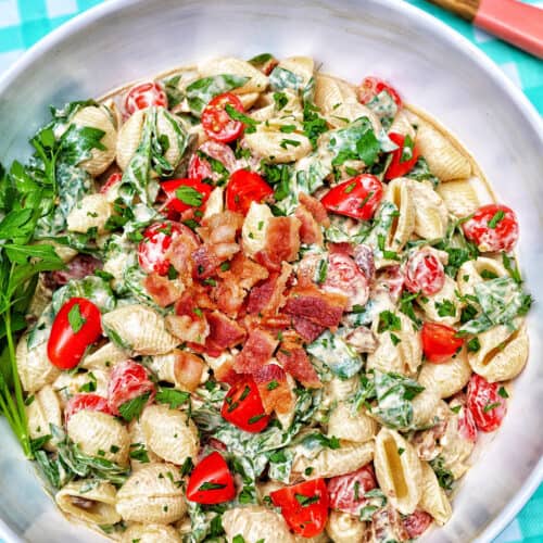 BLT pasta salad in a white bowl with wooden serving spoons overhead on an aqua checkered tablecloth.