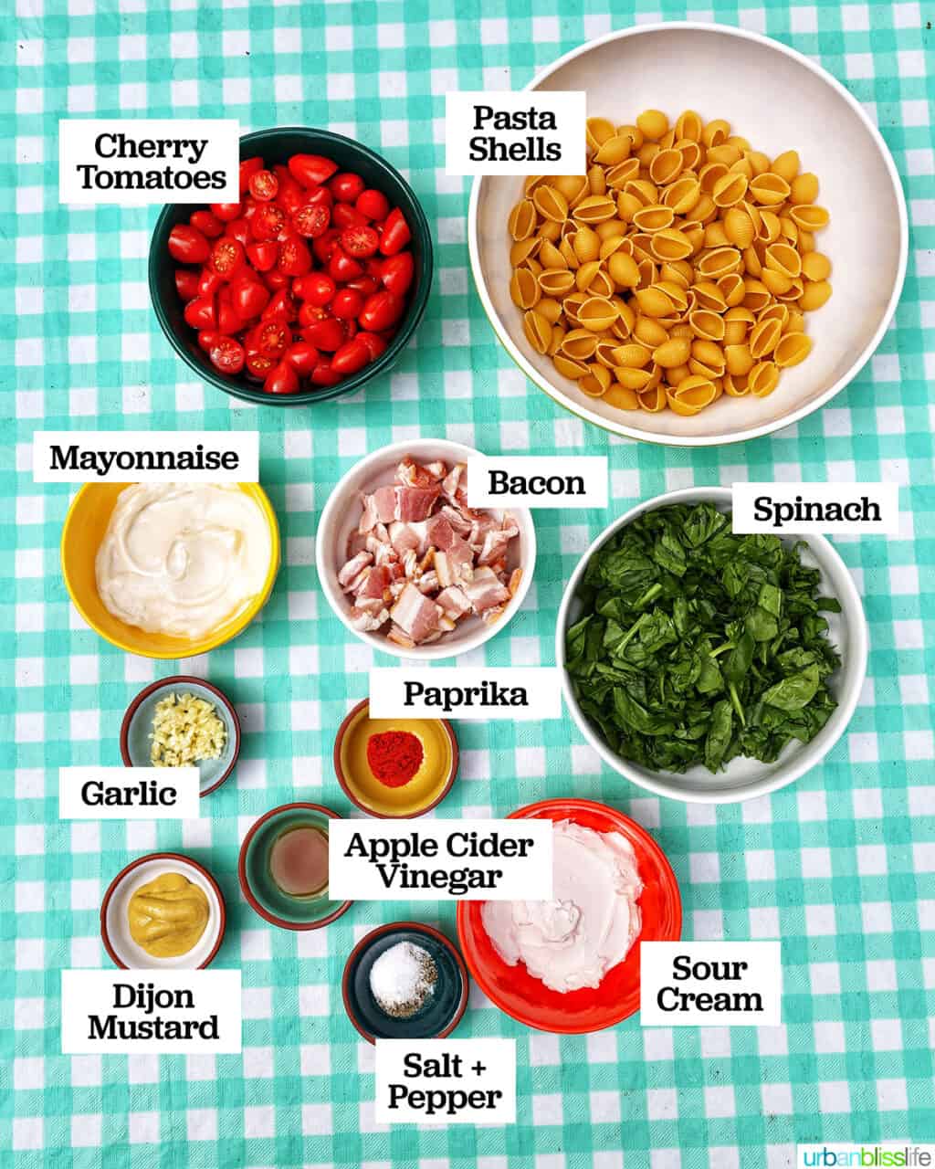 ingredients in bowls to make BLT pasta salad on an aqua checkered tablecloth.