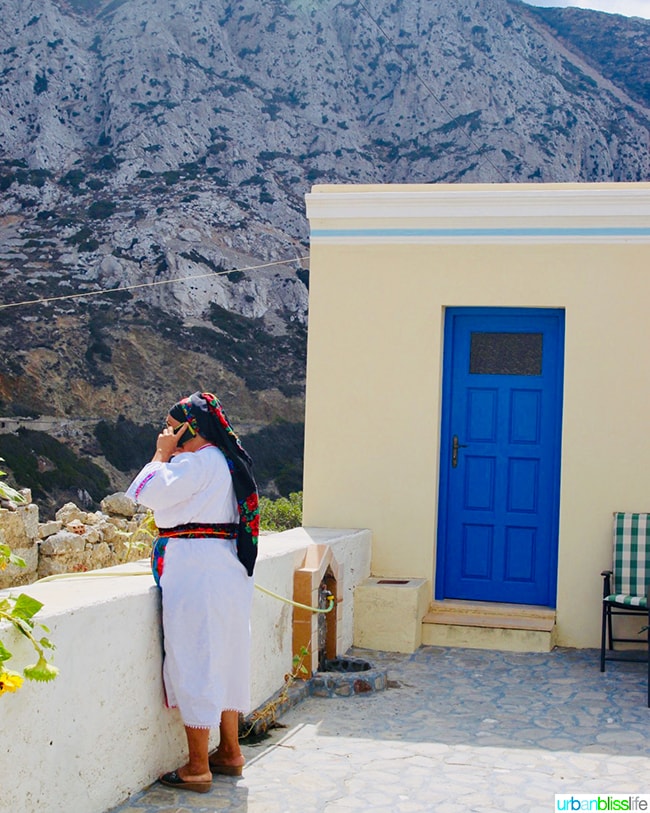 Karpathian woman dressed in traditional Greek dress on phone next to a home with bright blue door on Karpathos, Greece.