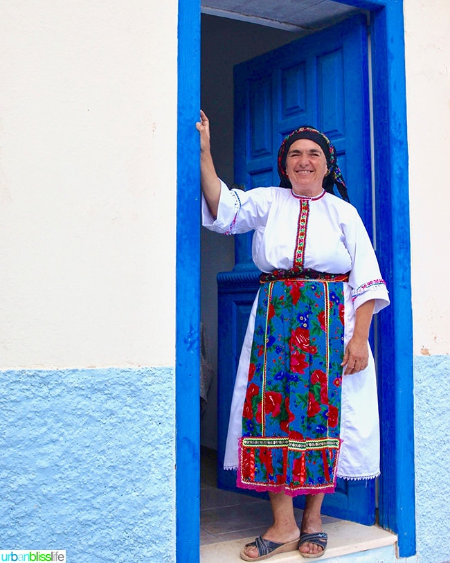 Karpathian woman dressed in traditional white and floral scarf and dress standing in a bright blue doorway of a bright white house in Olympos village, Karpathos island, Greece.