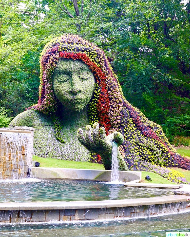 garden sculpture of a goddess with multcolored flowers for her hair, behind a water fountain.