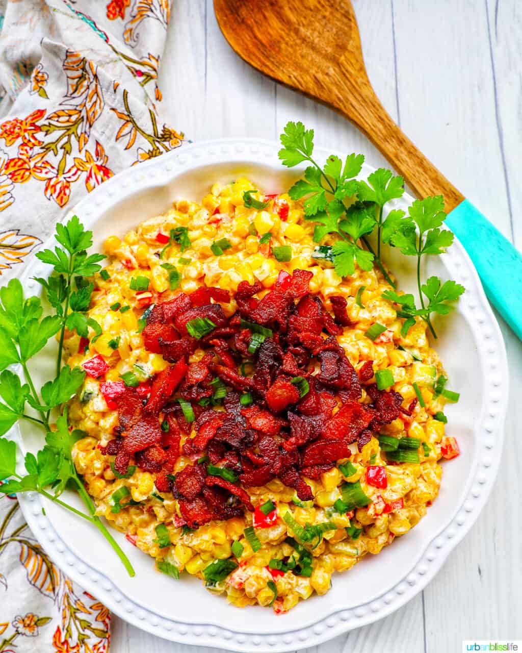 creamy confetti corn with crispy bacon, green onions, peppers, in a white bowl with greens and wooden spoon.