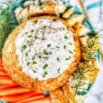 smoked salmon artichoke dip in a bread bowl with chives and rosemary garnish served with bread and carrots.