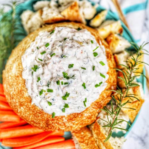 smoked salmon artichoke dip in a bread bowl with chives and rosemary garnish served with bread and carrots.