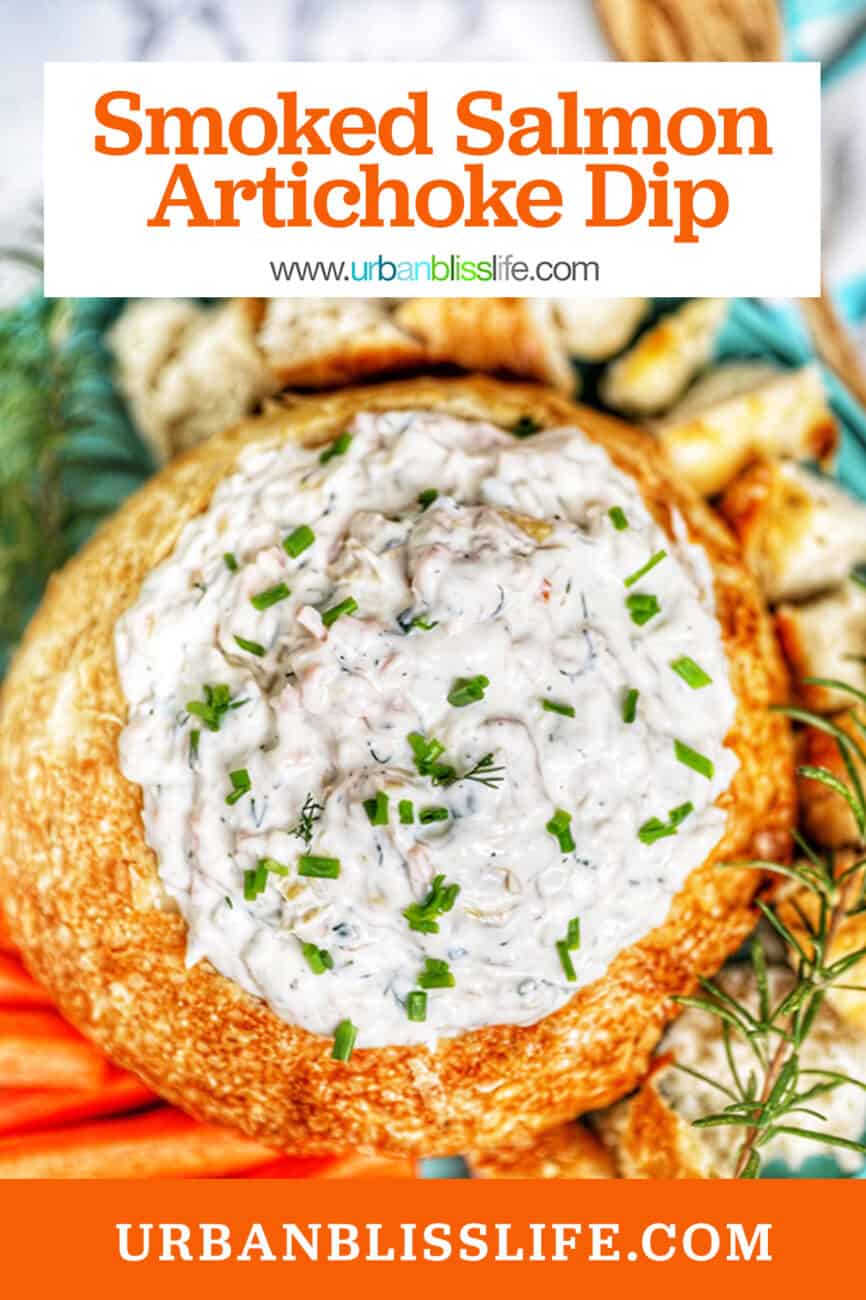 smoked salmon artichoke dip in a bread bowl with chives and rosemary garnish served with bread and carrots.