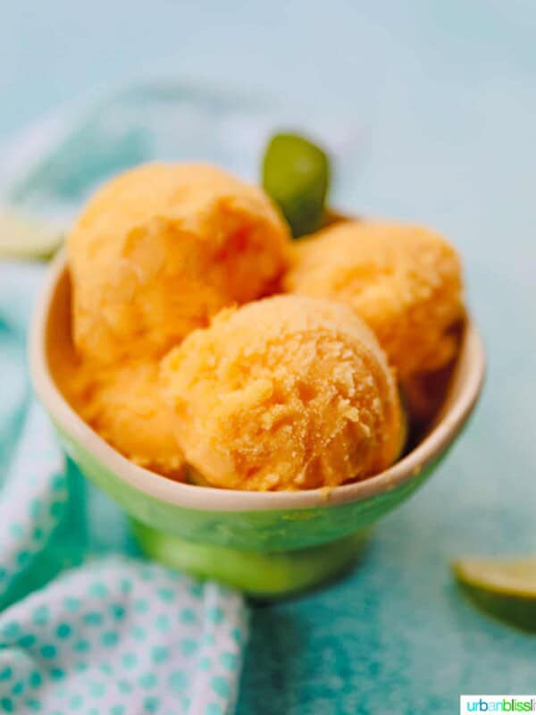 bowl of scoops of pineapple mango sorbet on a bright blue background with napkin and limes on the side.