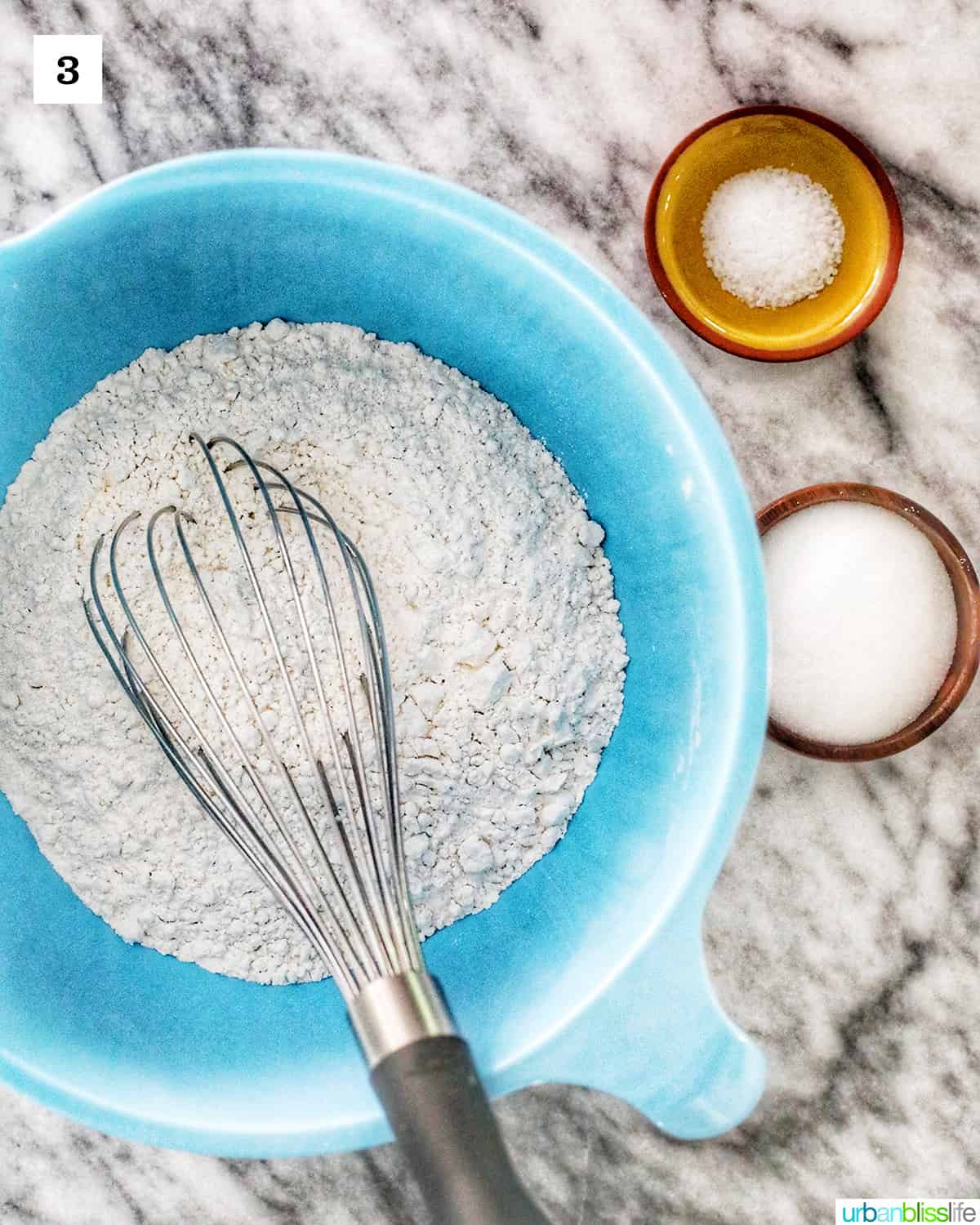 bowls of dry ingredients on a marble table, with whisk.