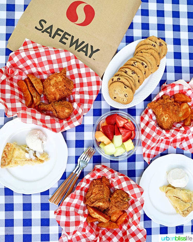 summer outdoor picnic with fried chicken, jo jos, cookies, fruit, on a blue and white checkered tablecloth