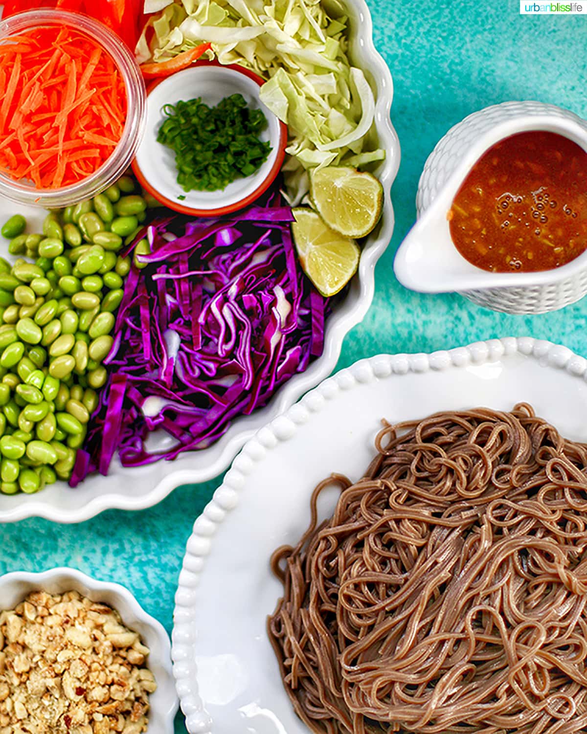 bowls of vegetables and soba noodles with a side pitcher of peanut sauce on a blue table.