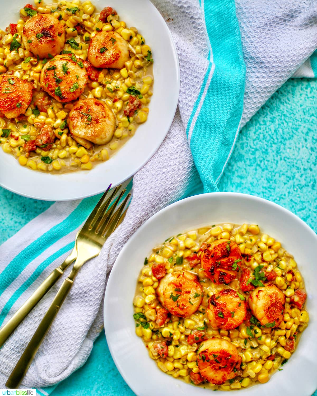 two bowls of seared scallops with corn and longganisa in a white bowl with two forks, a napkin, on a bright blue background.