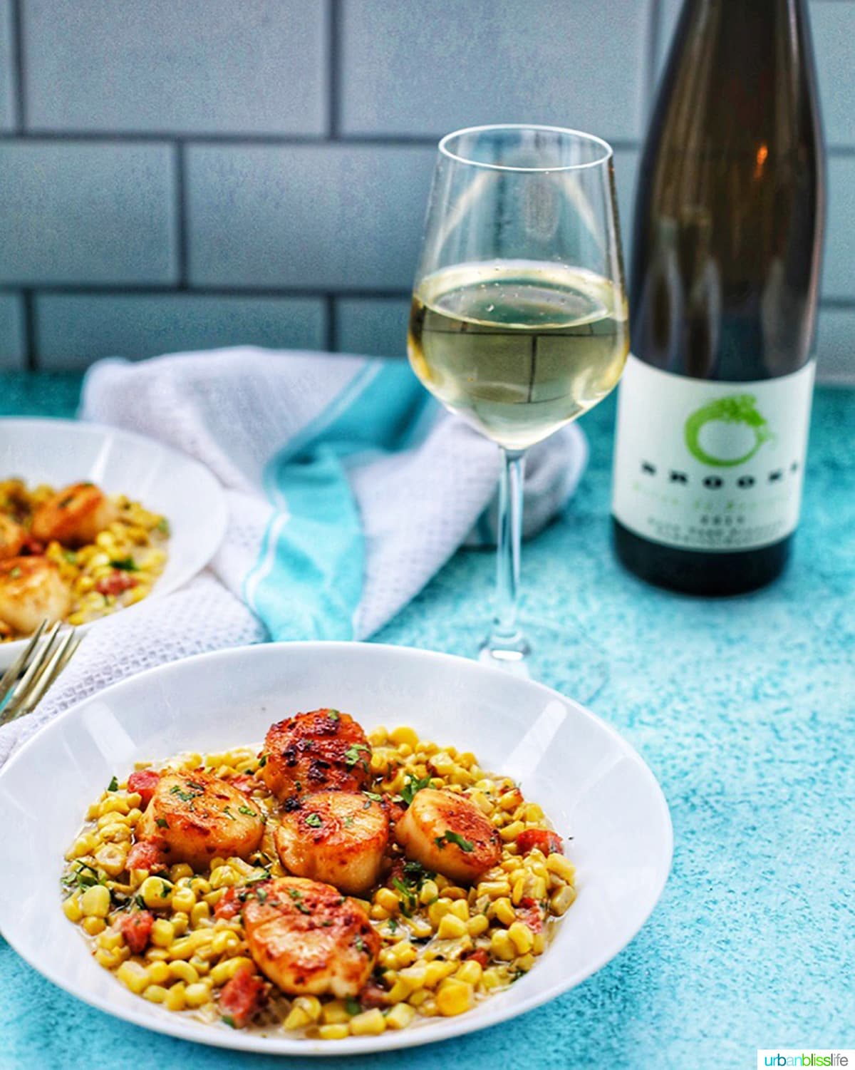 Seared scallops with corn and longganisa in a white bowl with white wine in a glass and bottle on a blue background.