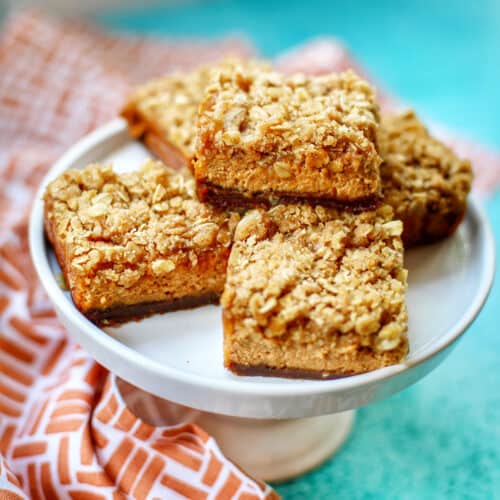 pumpkin cheesecake bars on a white cake stand with kitchen towel