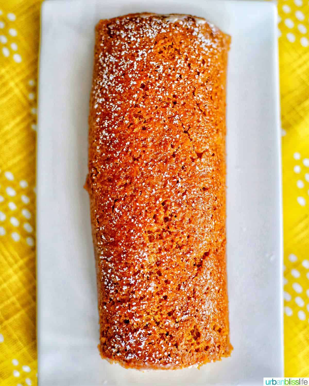 pumpkin roll with cream cheese filling uncut on a white plate and yellow tablecloth.