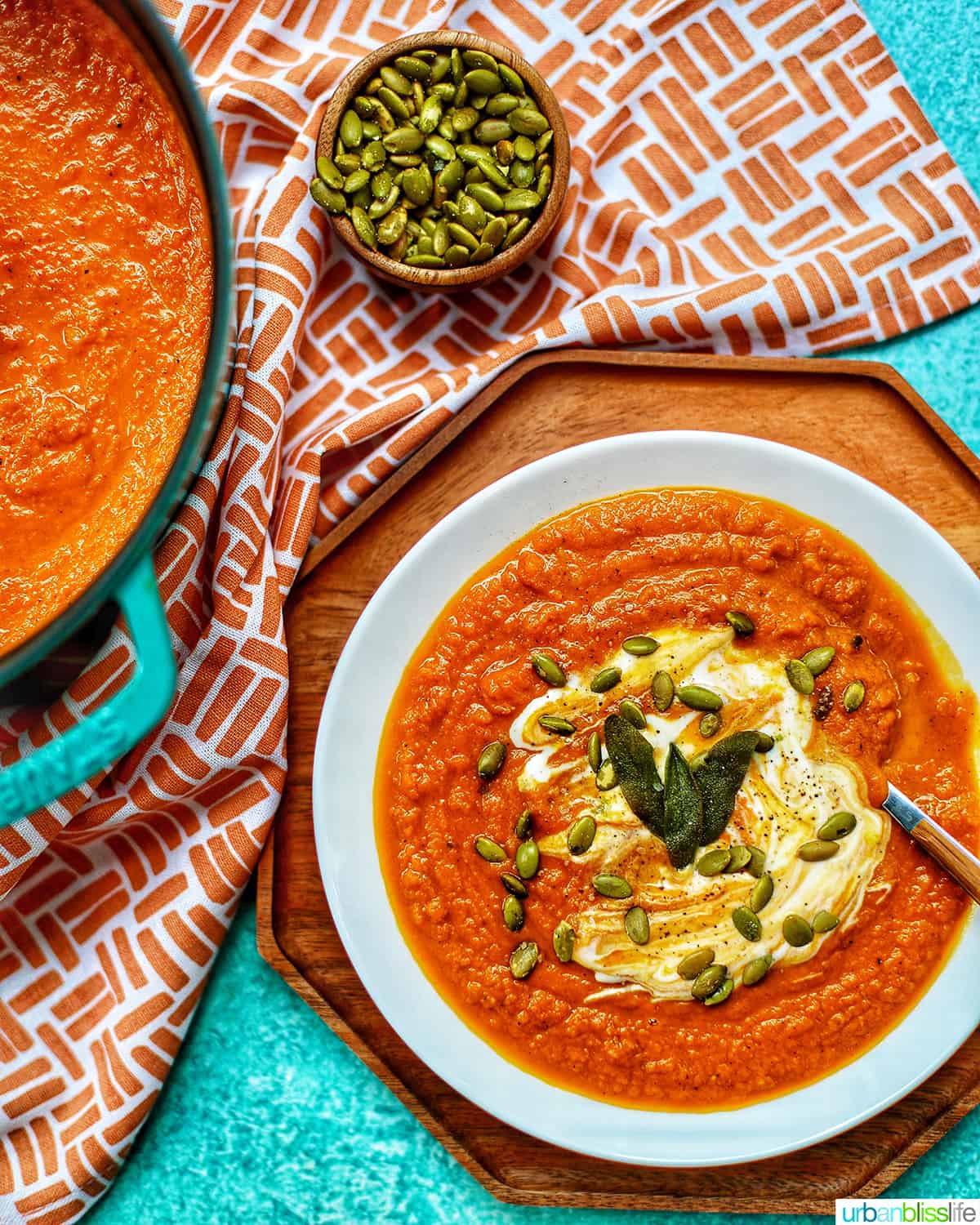 white bowl of vegan pumpkin soup on top of a wood tray next to dutch oven filled with pumpkin soup on bright blue background with orange and white napkin.