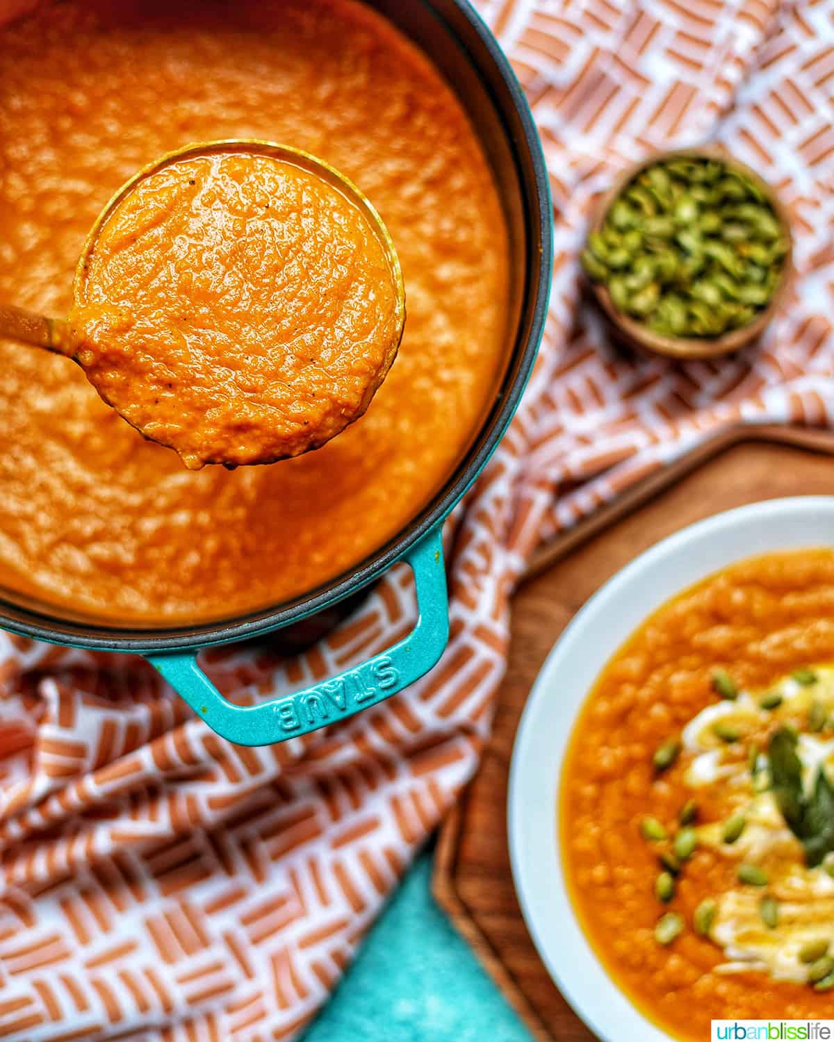 vegan pumpkin soup in ladle above a dutch oven of pumpkin soup without cream, next to bowls of soup and pumpkin seeds.