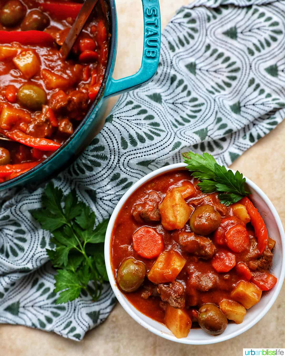 bowl and large blue Staub pot of of beef caldereta with potatoes, green olives, red peppers, tomatoes, on green and white napkin.