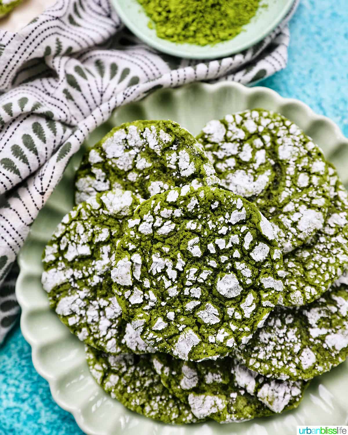 plate of bright green matcha crinkle cookies stacked on top of one another on a bright blue background.