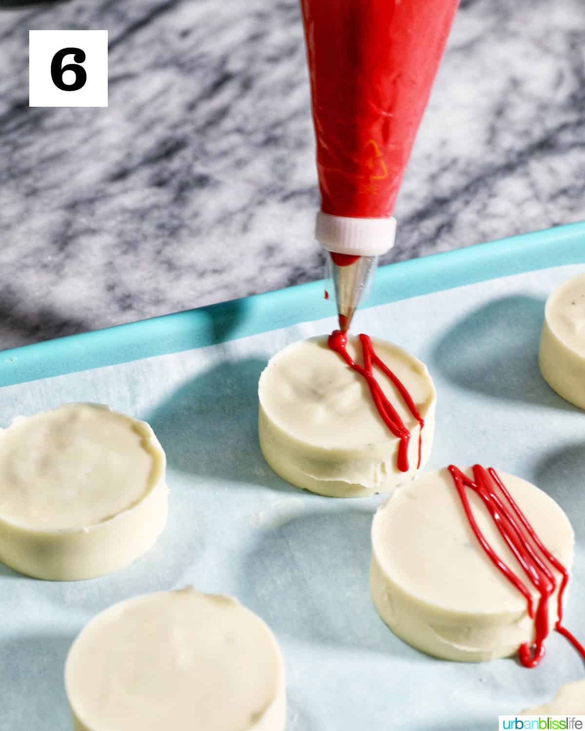 red frosting being piped onto white chocolate covered Oreo cookies on parchment paper lined blue baking sheet.