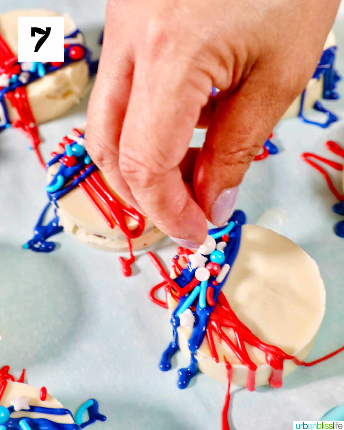 Hand sprinkling red, white, and blue sprinkles onto frosting of 4th of July Oreos.