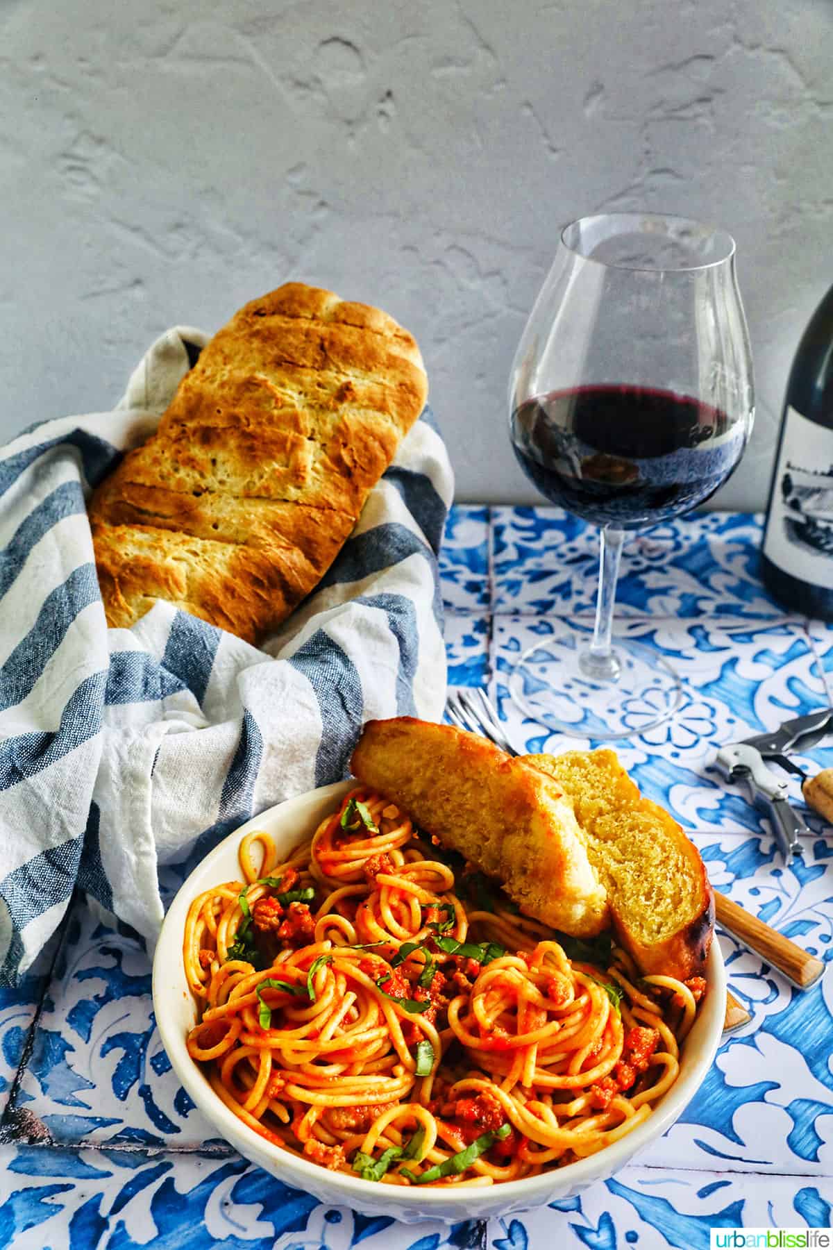 bowl of spaghetti, side of french bread in blue and white towel, glass of red wine, all on a tiled table.