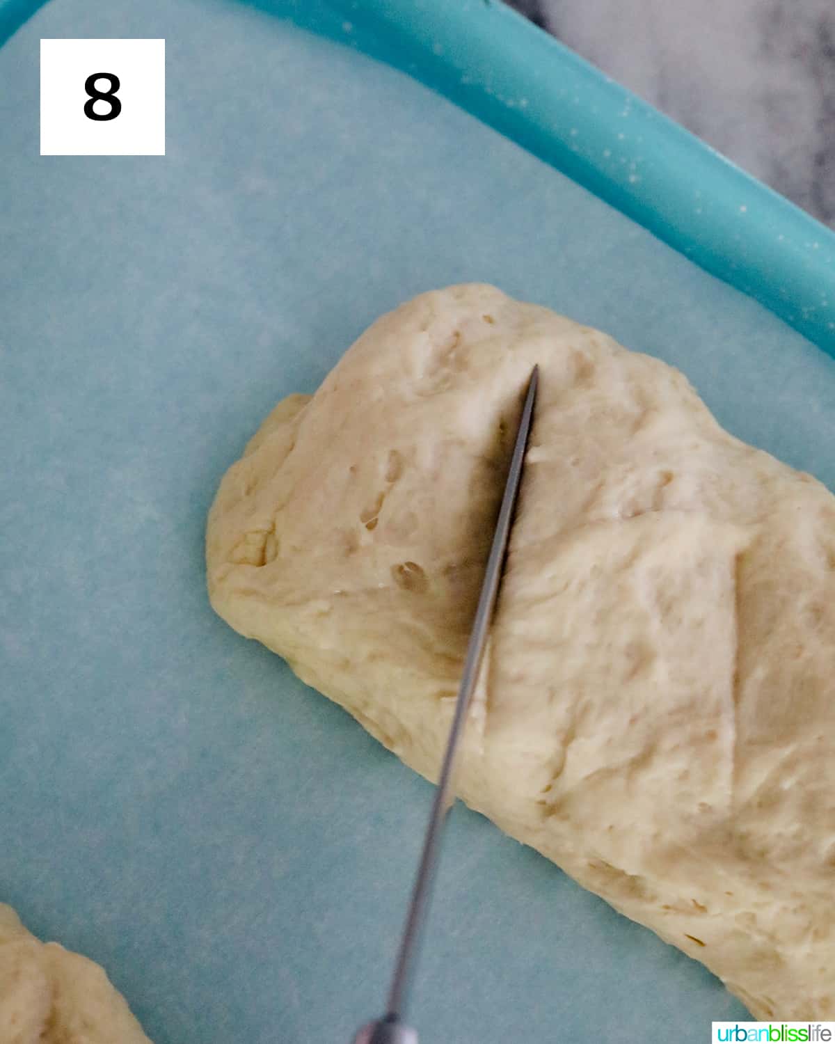 a knife scoring a raw french bread loaf on a parchment paper-lined baking sheet.