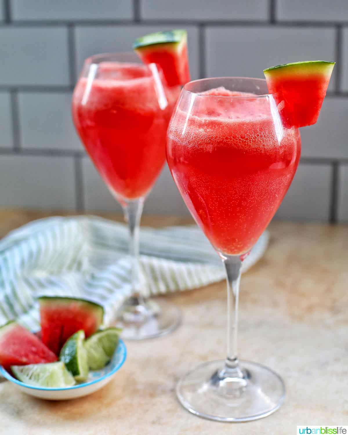 two glasses of watermelon mimosas with bottle of sparkling wine and watermelon slices in the background.