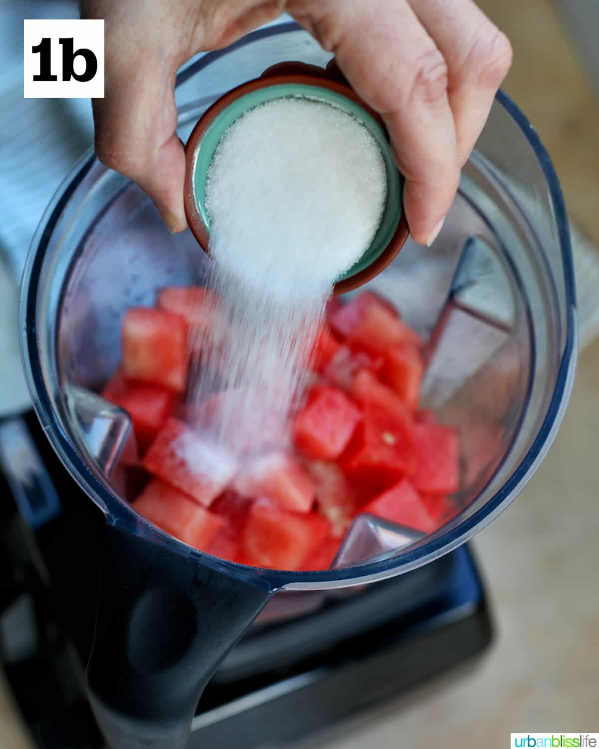 hand adding sugar in a small bowl into a blender with cubed watermelons.