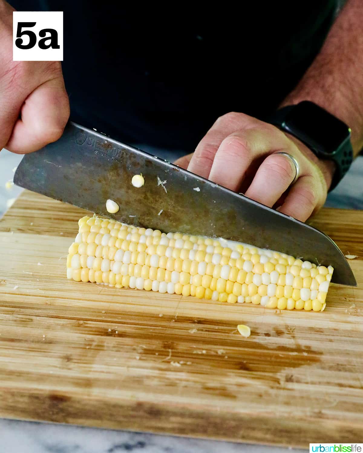 hands using a knife to slice down a halved corn on the cob on a cutting board.