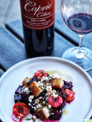 bottle and glass of red wine with a colorful beet, cherry, pork belly salad on a white plate.
