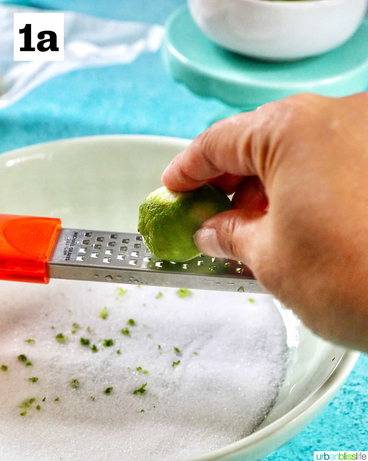 hand zesting a key lime over a bowl of granulated sugar.