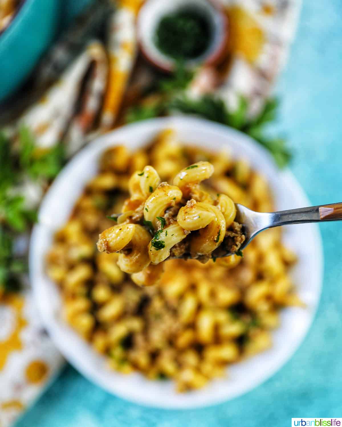 forkful of dairy free cheeseburger pasta above a bowl of cheeseburger pasta on a blue background.