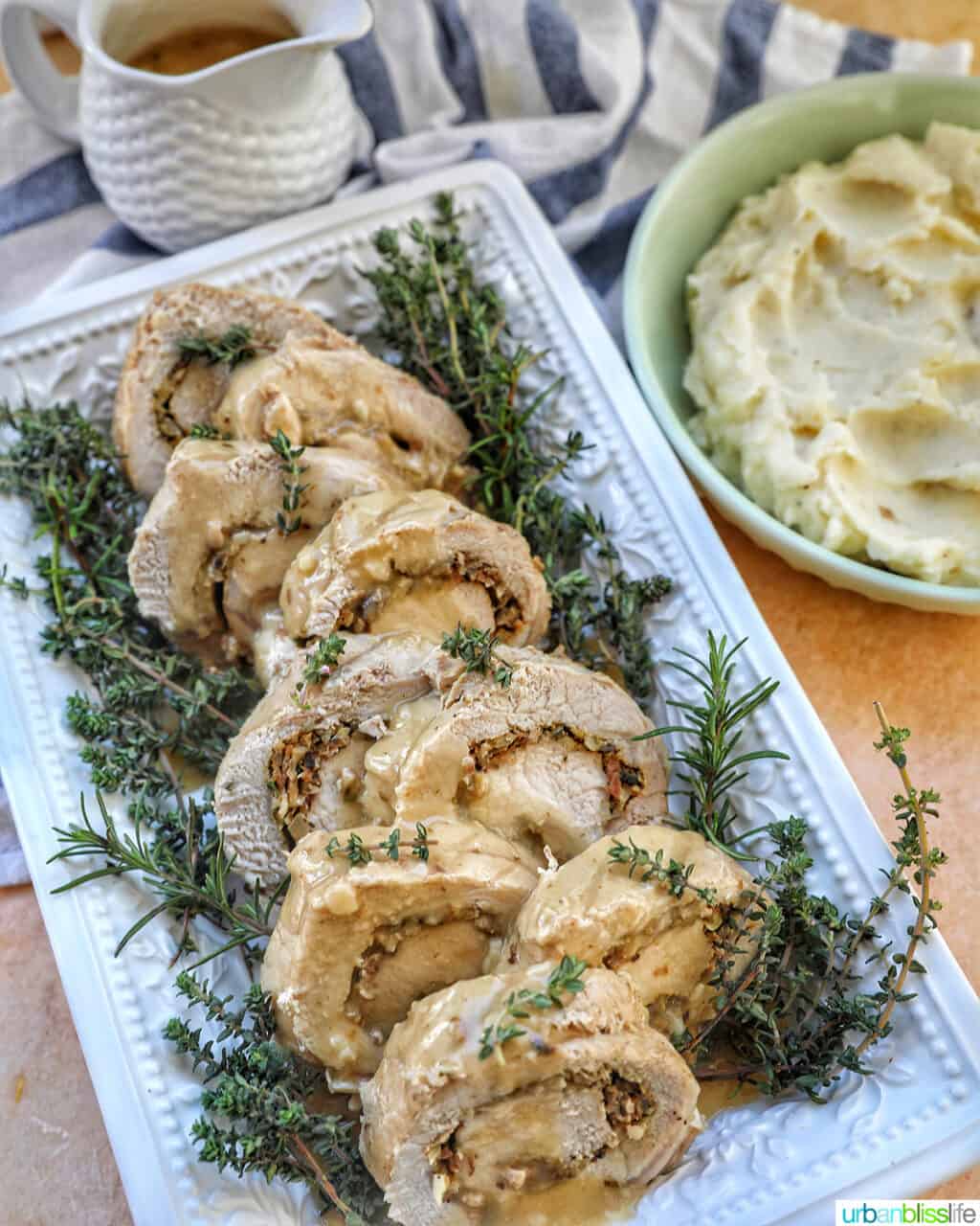 sliced stuffed pork loin on a white plate with a bed of herbs with side of mashed potatoes and gravy.