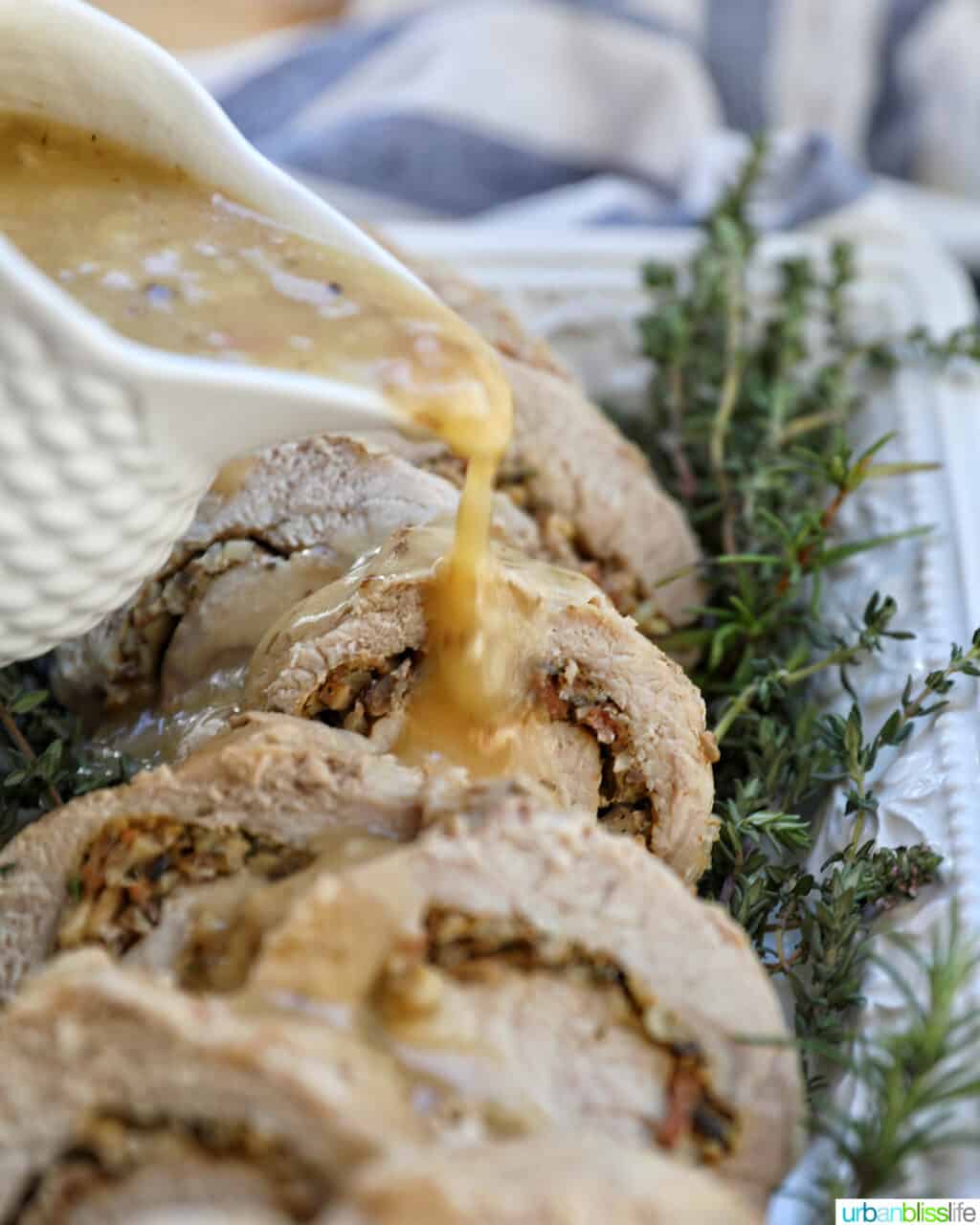 pitcher of gravy pouring over sliced stuffed pork loin on a white plate with herbs.