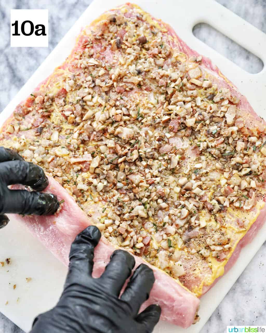 gloved hands rolling up the stuffed pork loin on a white cutting board.