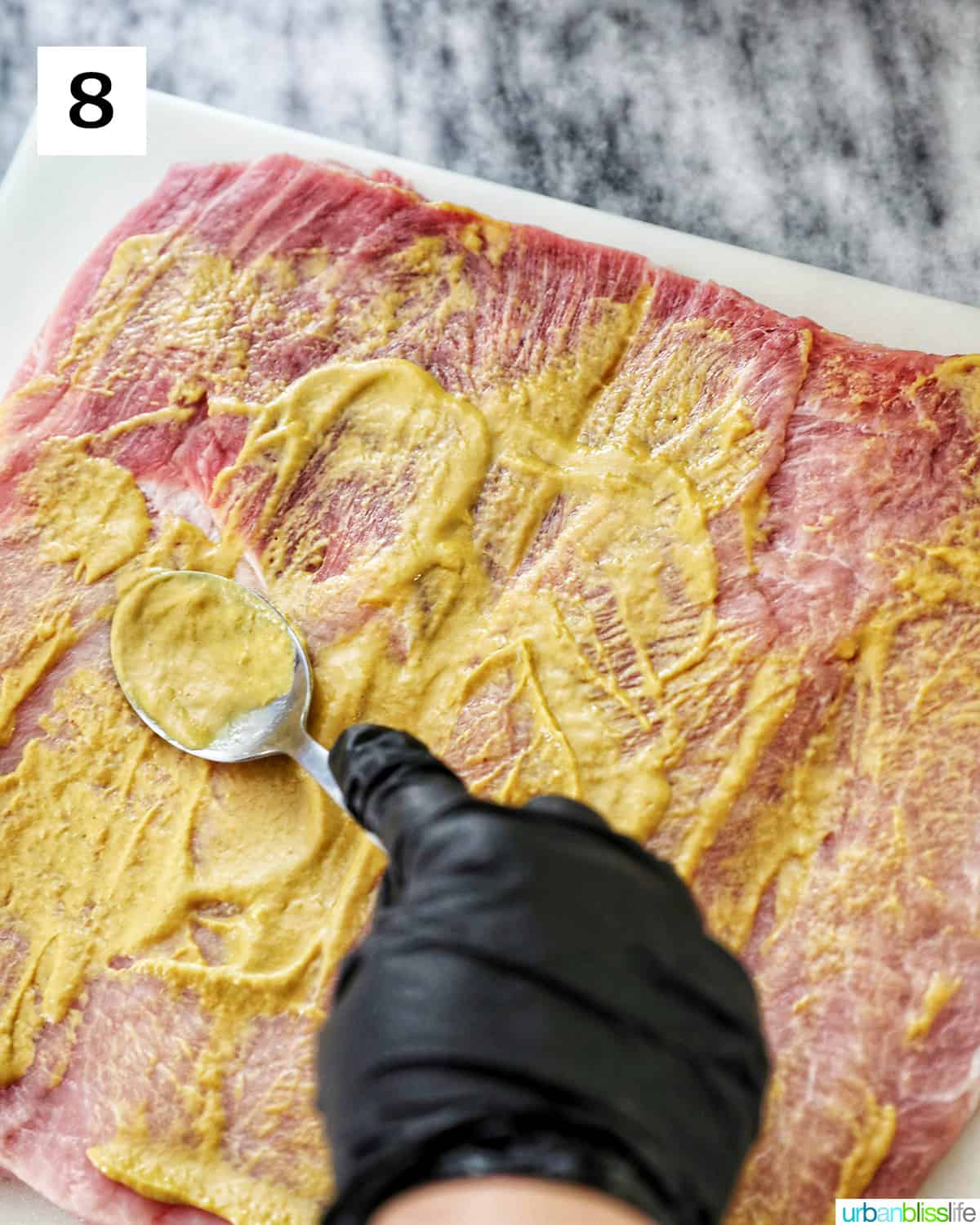 gloved hand spooning mustard over flattened pork loin.